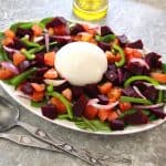 An oval platter with a beets salad with burrata in the middle. Serving utensils are in front of the platter and an olive oil dispenser is on the back.