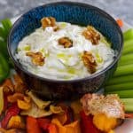 A bowl of sour cream dip in the middle of chips and vegetables. Dip is garnished with walnuts and olive oil.