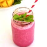 Mango Dragon Fruit Smoothie shown with some fresh fruits in the background, a striped straw and fresh mint leaves