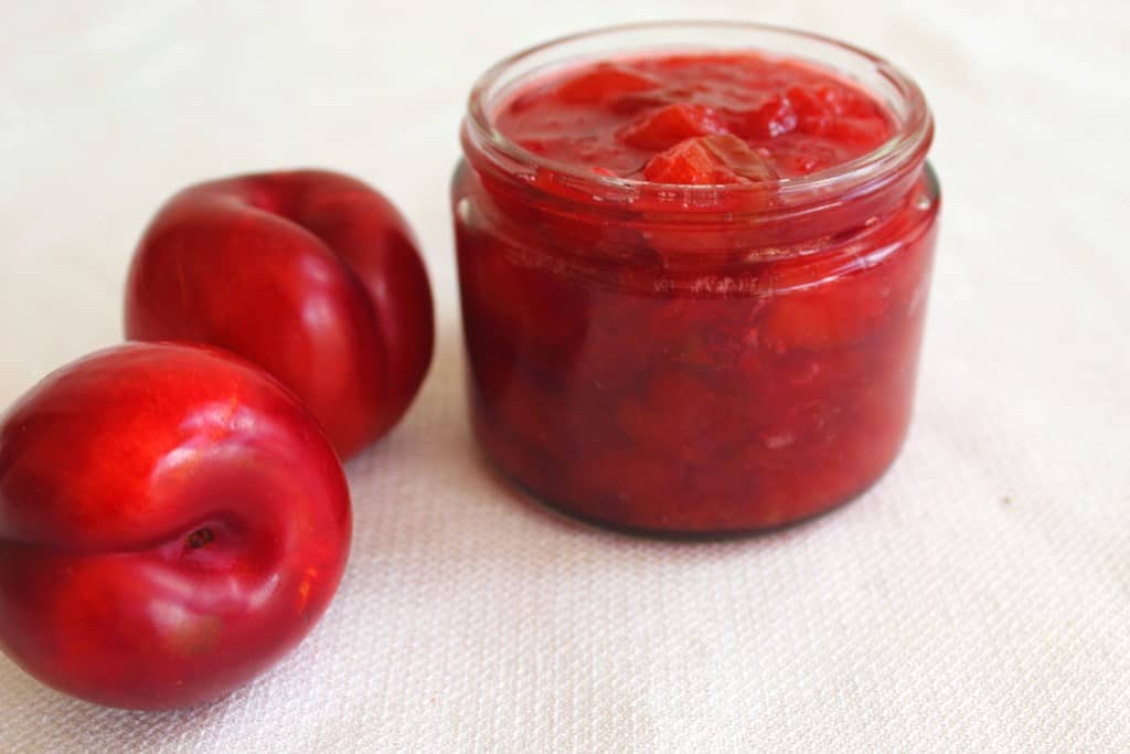 Simple, homemade Red Plum Jam with no pectin. Picture shows 1 jar of jam and 2 fresh plums. In Albanian, plum jam is called  reçel kumbulle.