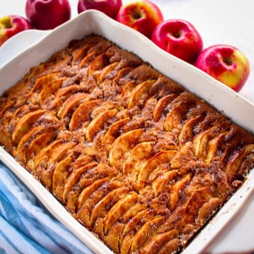 An apple cake on a baking dish, cake is topped with apple slices. Next to the baking dish there are fresh apples, red and crisp looking.