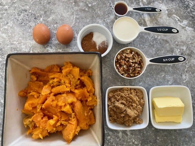 Ingredients for filling layed out: scooped out sweet potatoes in a bowl, brown sugar, butter, chopped pecans, cinnamon, nutmeg, salt, eggs, heavy cream and vanilla.