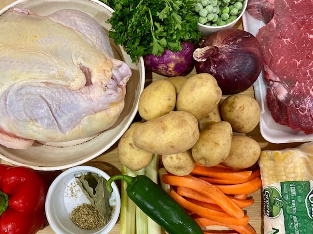 Picture shows most of the ingredients to make Christmas Soup - whole chicken, beef, onion, turnip, parsley, potatoes, corn, red bell pepper, oregano, salt, bay leaves and carrots. Missing from the picture are olive oil and tunta.