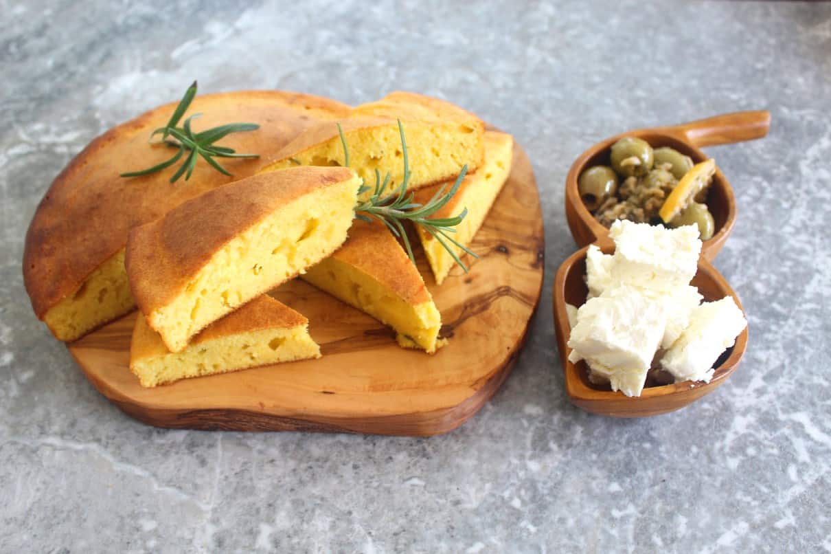 Rosemary cornbread is served sliced on a wood board and decorated with rosemary twigs. Next to it there are two wooden round bowls filled with chunks of feta cheese and olives.