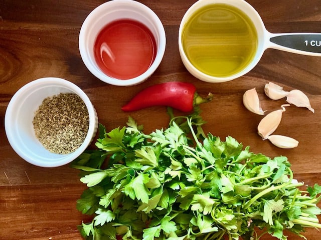 Ingredients to make homemade chimichurri in a cutting board: parsley, oregano, red wine vinegar, olive oil, red chili pepper and garlic. 