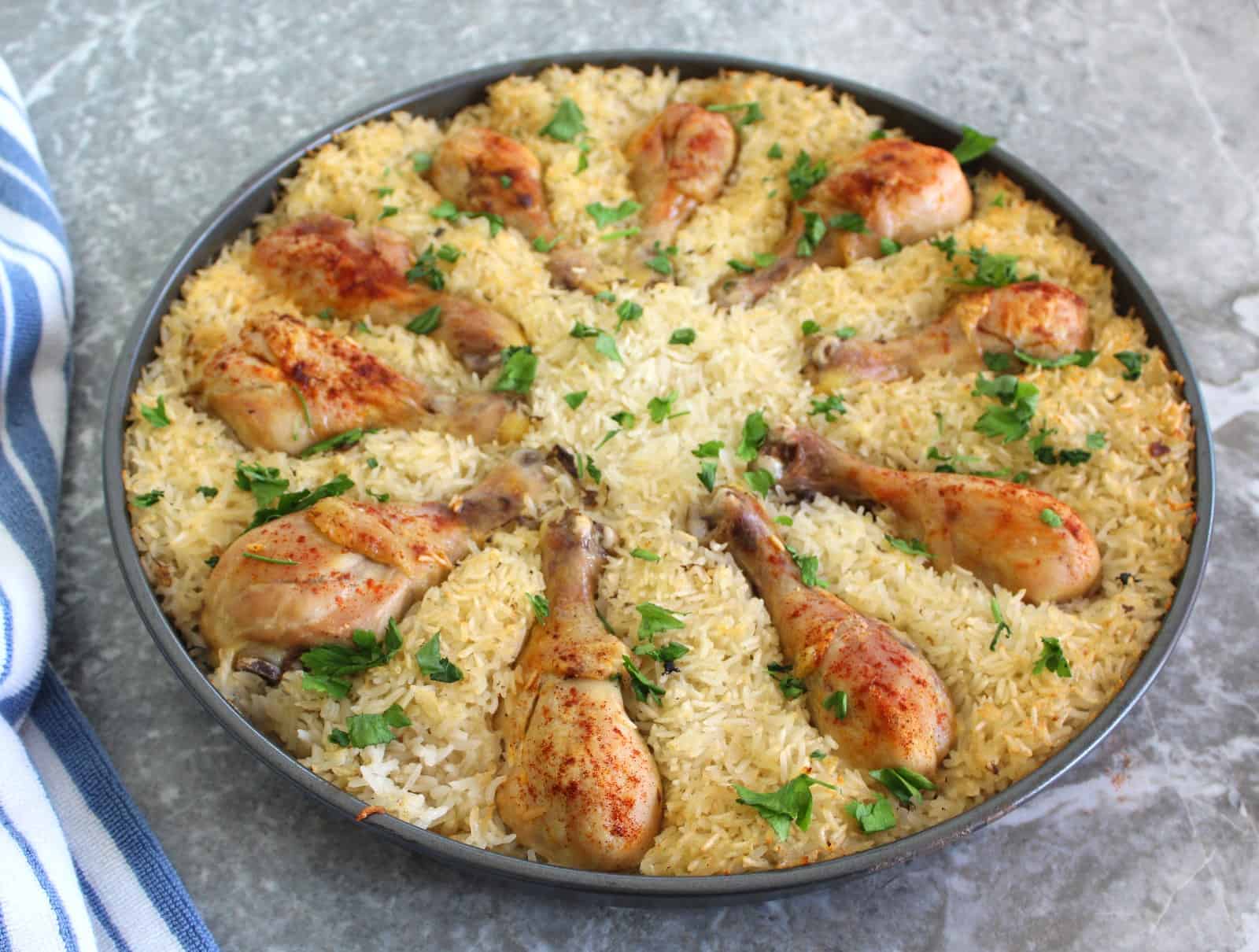 Round tray with a chicken & rice dish garnished with chopped parsley. Chicken drumsticks are also laid out in a circular shape.