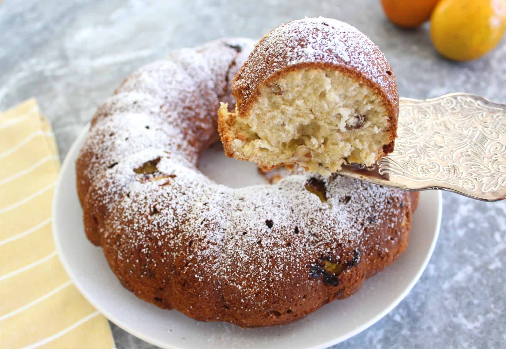 Orange bundt cake served in a white plate, shown with a slice being lifted with a silver spatula from the plate. 