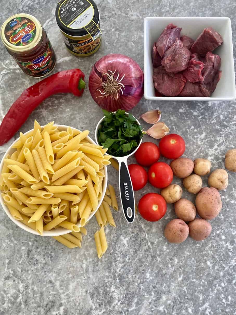 Ingredients to make the pasta dish all layed out, ready to cook: Aji Panca, Bouillon, red pepper, red onion, parsley, garlic, meat chunks, penne riggati, tomatoes, potatoes.