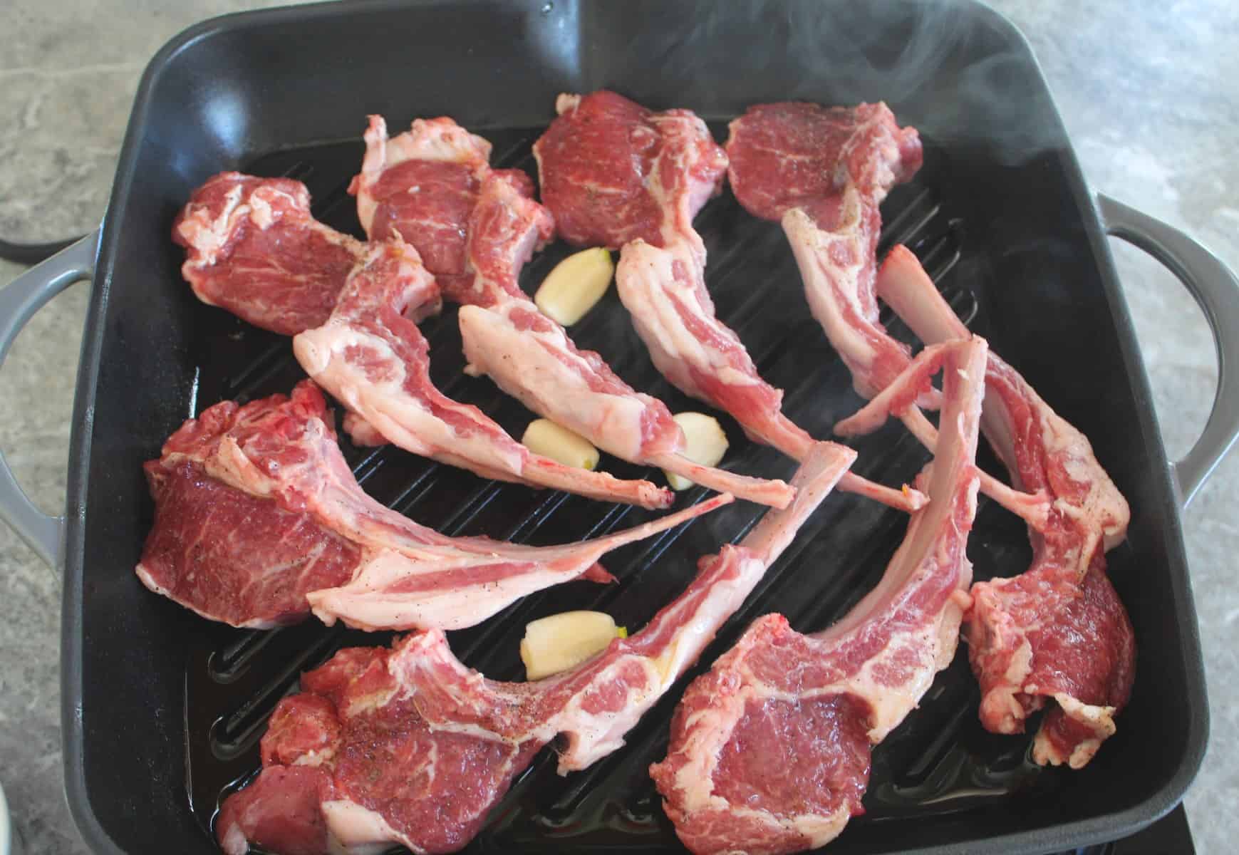 Lamb chops in the cast iron grill pan with garlic being seared on one side. The top side is not grilled yet. The picture even captures some smoke & steam coming out of the pan. 