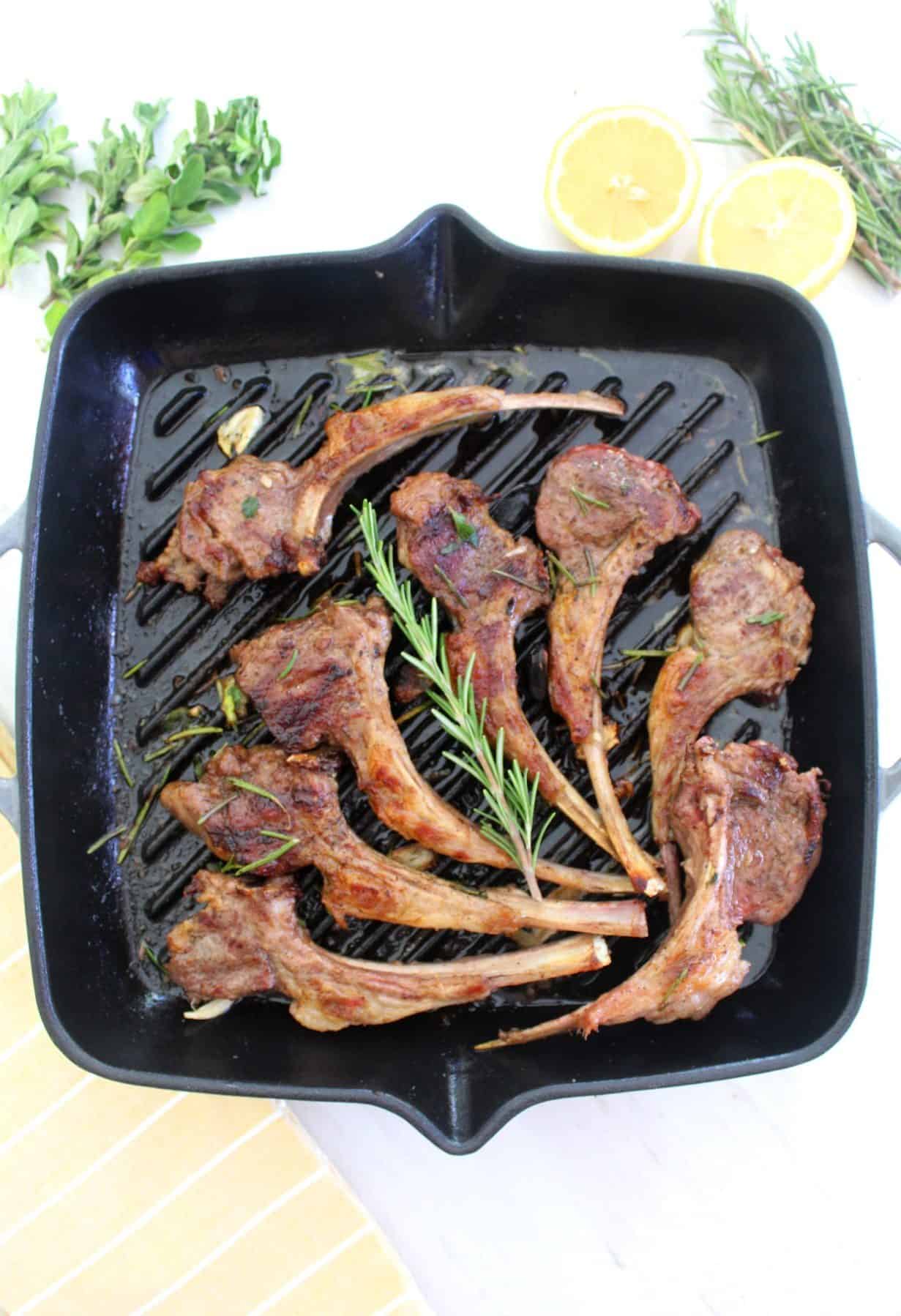 Lamb chops on cast iron pan, decorated with rosemary. Picture is styled with some fresh lemon, rosemary and oregano on the side of the pan.