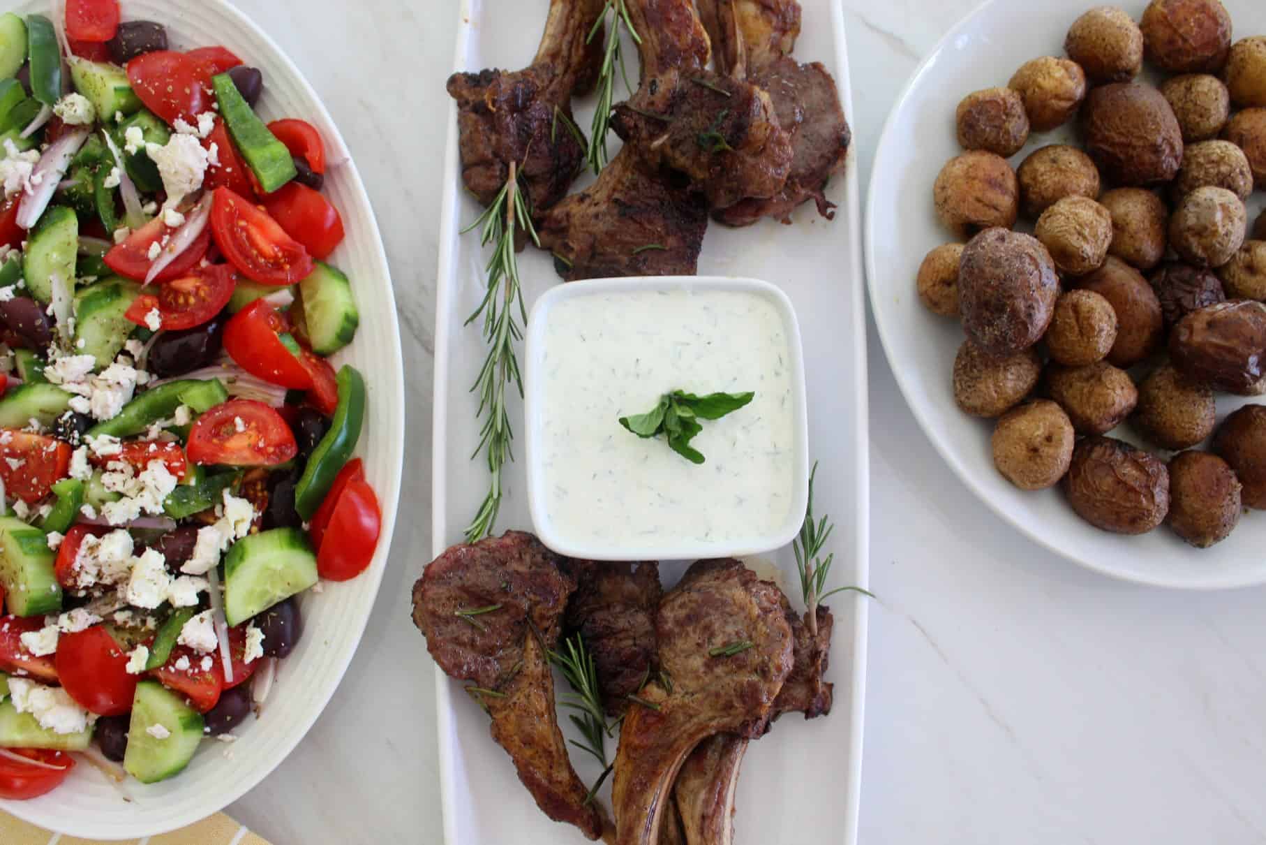 A platter of lamb chops, about 4 on each side with a tzatziki dip in the middle. On one side of this platter, you see a Mediterranean salad and on the other side you see roasted potatoes.  