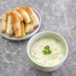 Fried yucca in a plate next to a bowl of a yogurt based dip. Dip is garnished with cilantro.