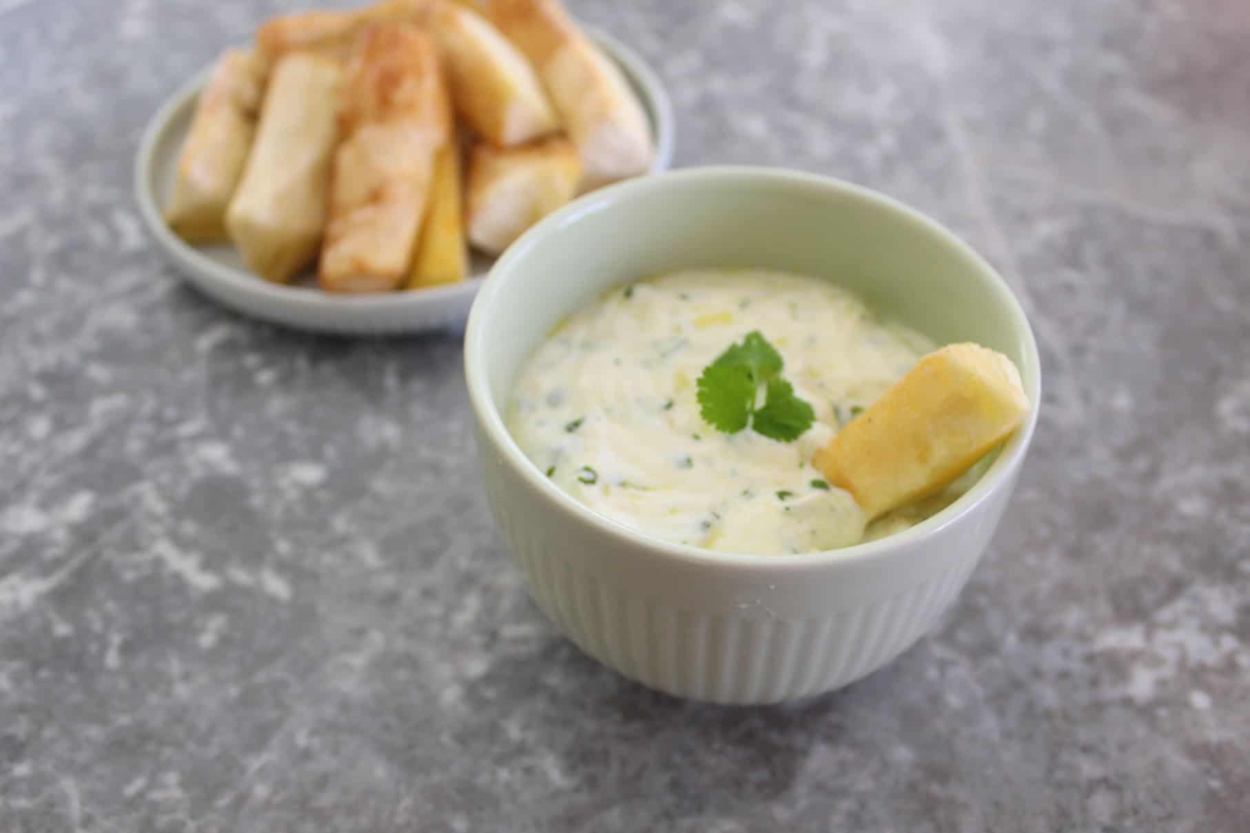 Yogurt based dip with cilantro, shown with fried yucca. One yucca stick is dipped in the dip while the plate of fried yucca is shown behind the dip.