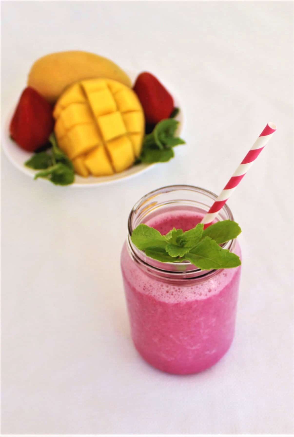 Tropical, Summer Mango Dragon Fruit Smoothie - Smoothie is shown with a straw and mint leaves in the forefront and fresh fruits in the background.