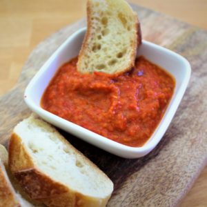Red pepper dip shown in a white recipient with bread dipping in it.