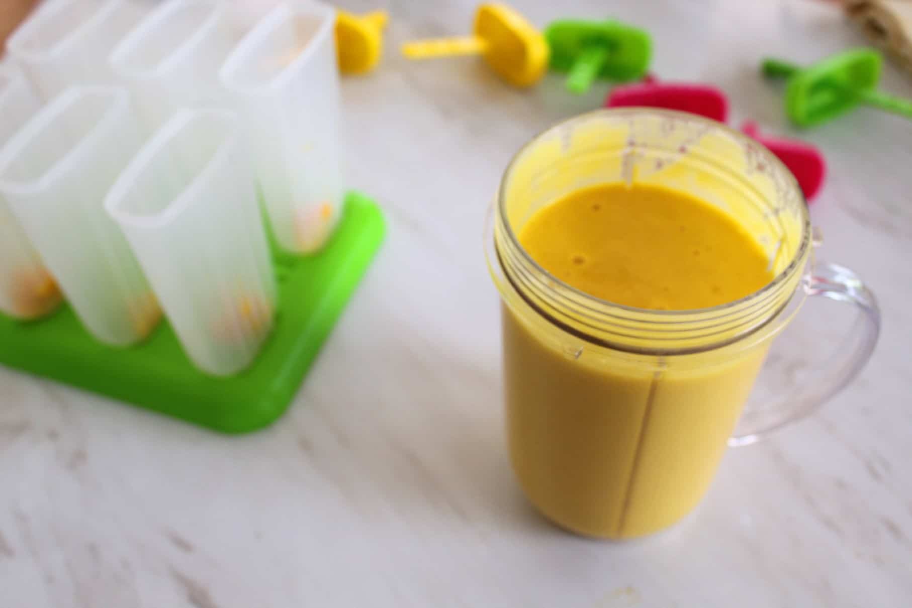 Blended mango mix with coconut milk in a blender next to popsicle molds.