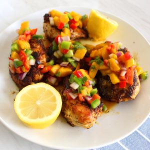 A plate with cooked chicken pieces, topped with mango salsa and garnished with 2 lemon halves.