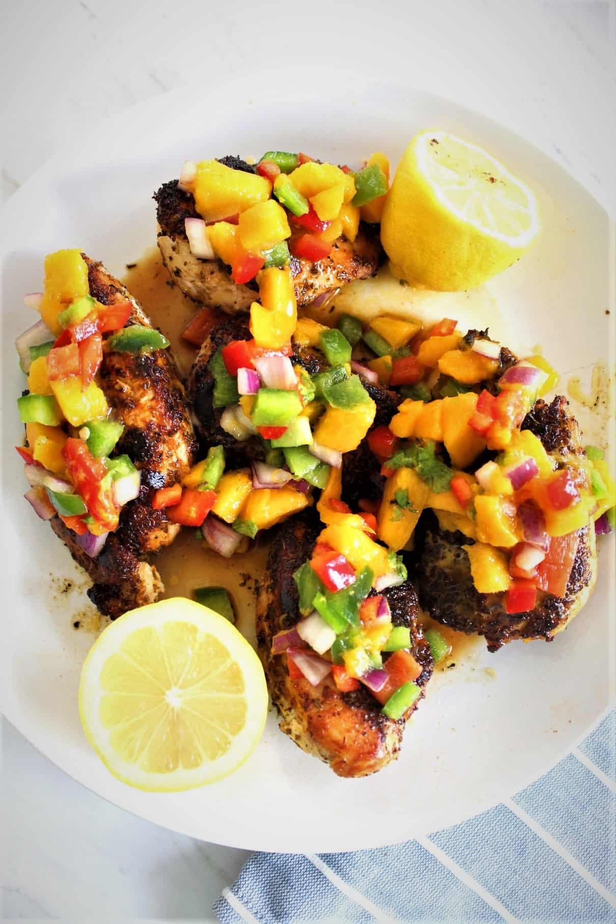 Skillet Chicken in Mango Salsa - picture shows a large round plate with cooked chicken topped with a mango salsa. There are 2 lemon halves in the plate too. 