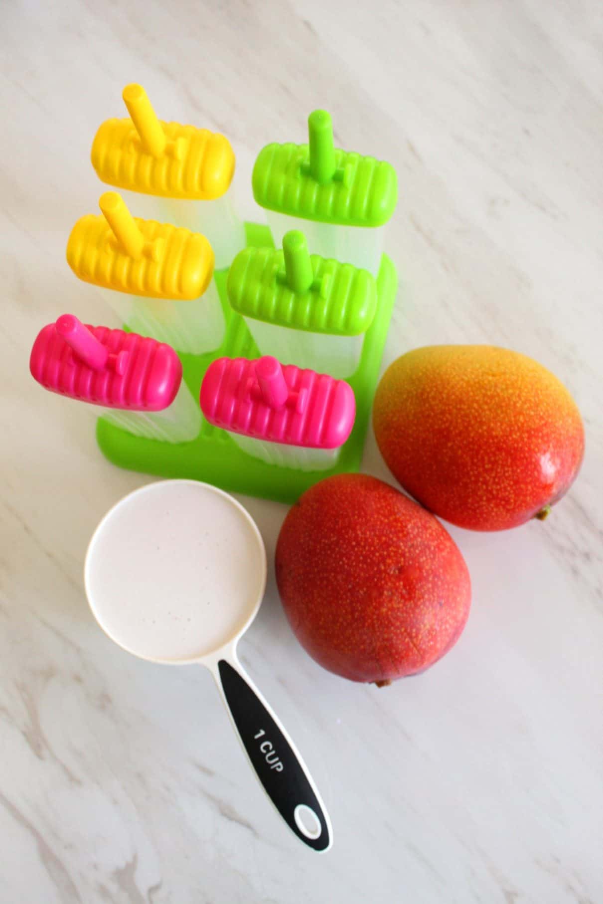 2 big size mangoes, a cup of coconut milks and molds for popsicles are laid out.