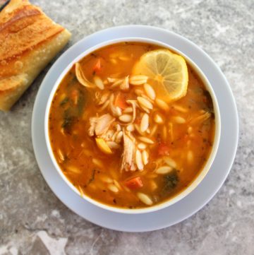 A bowl of orzo soup garnished with a slice of lemon shown with a piece of baguette next to the bowl, on the left hand side.