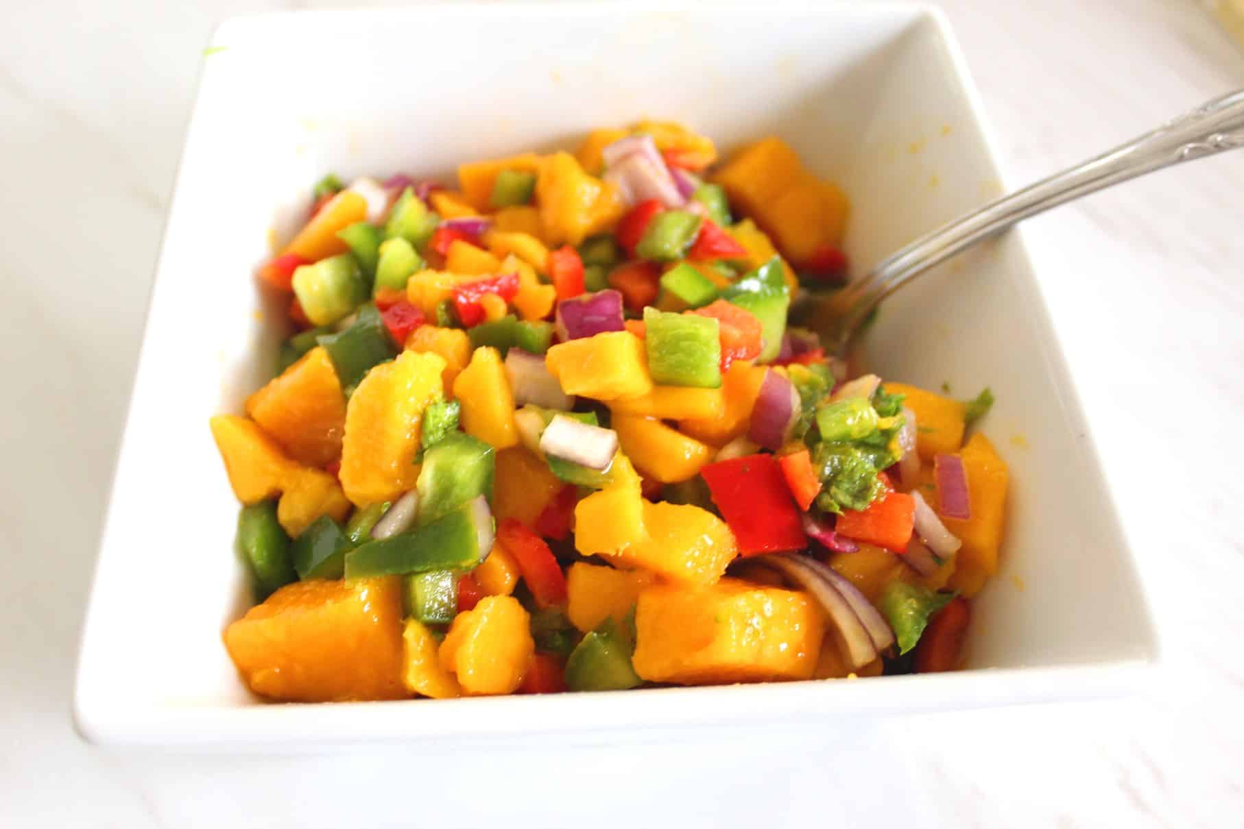 A square bowl with mango salsa that has a spoon in it. 