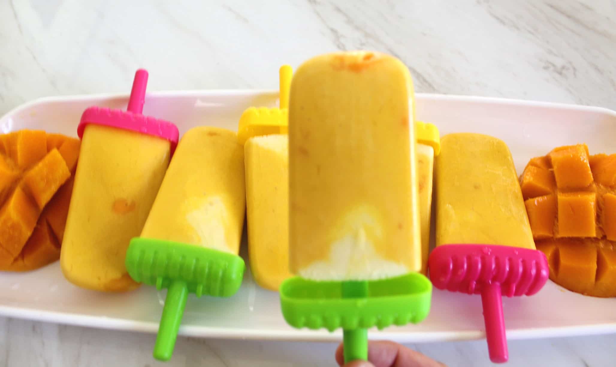 Yummy looking mango popsicles with different color handles laid out in a tray. There's fresh mangoes on both sides of the tray.