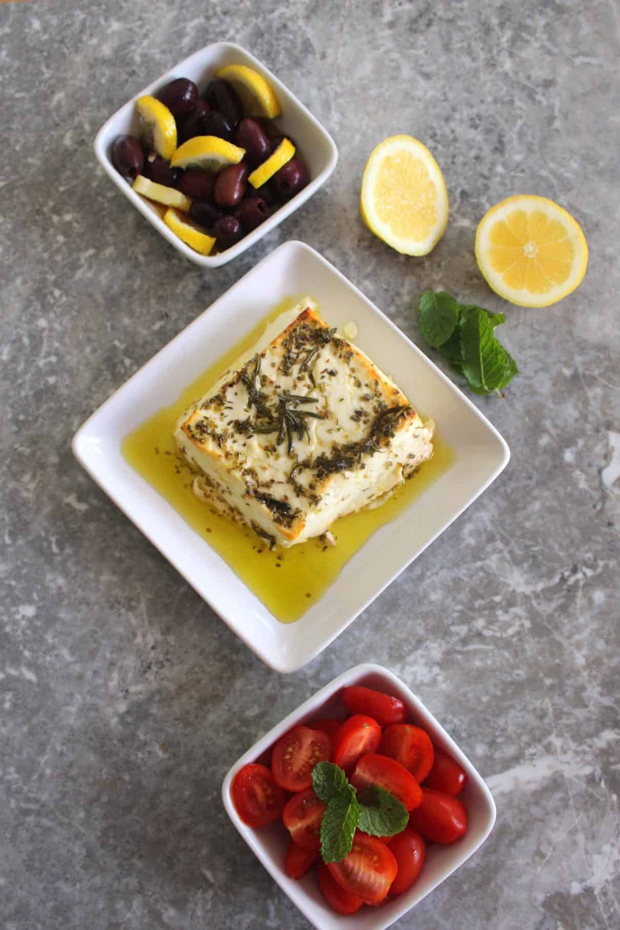 A set of appetizers: from top to bottom you see a dish with kalamata olives and lemon, then baked feta cheese with herbs and lastly grape tomatoes garnished with mint. Next to the 3 sides dishes there's lemon slices and mint leaves too.