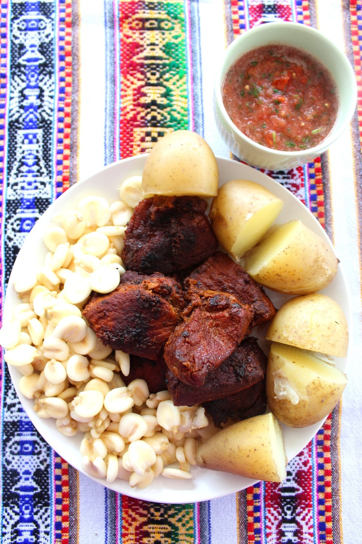 Pork chicharron served on a platter with mote (white large grain corn) and boiled potatoes. Next to the platter, there's a serving of a llajua (a Bolivian tomato salsa).