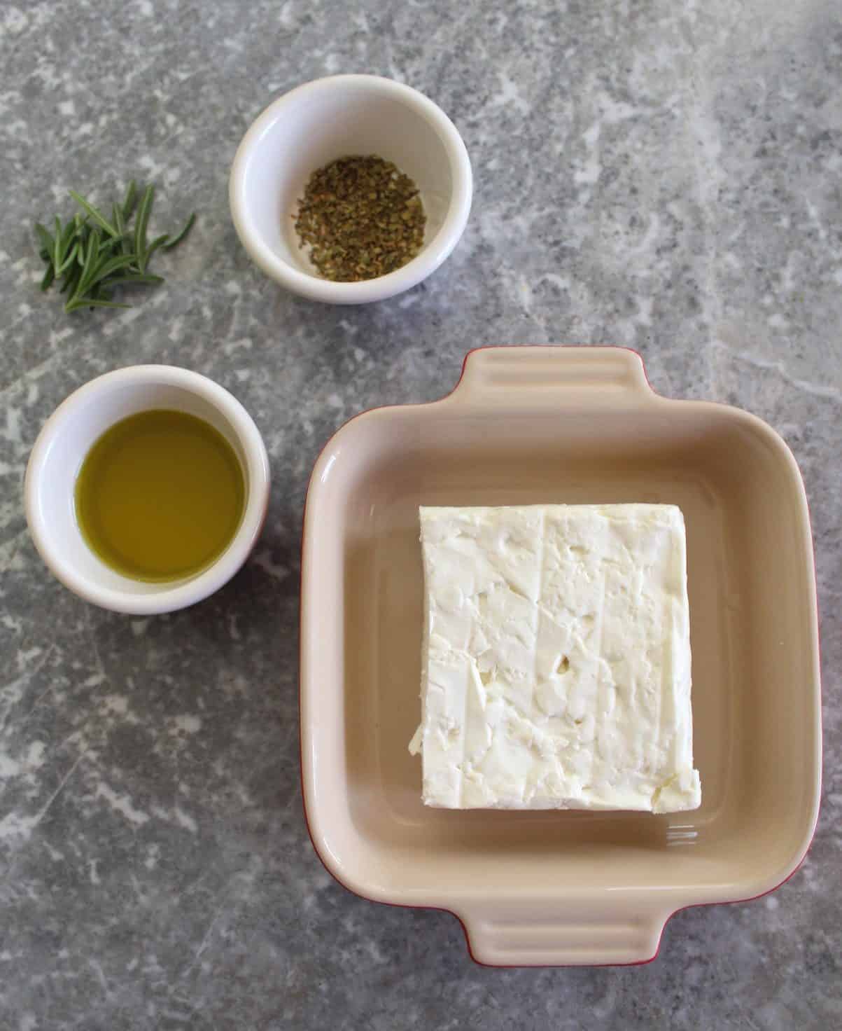 Ingredients for baked feta cheese - feta cheese block in a baking tray, fresh rosemary, dry oregano and olive oil.