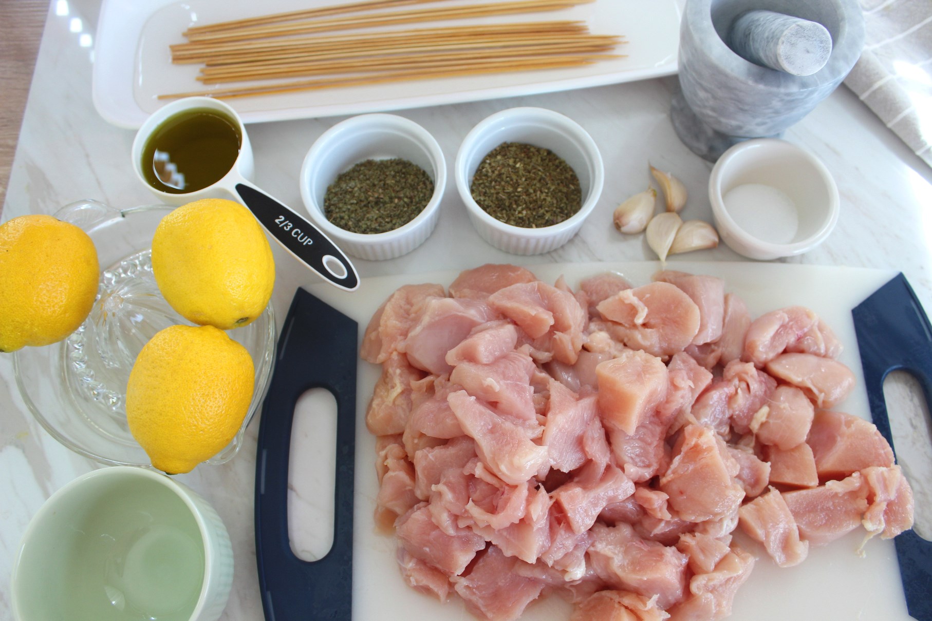 All the ingredients together on a flat surface for the chicken kabobs. You see herbs, lemons, cut chicken, a mortar and pestle, oil and skewers in water. 