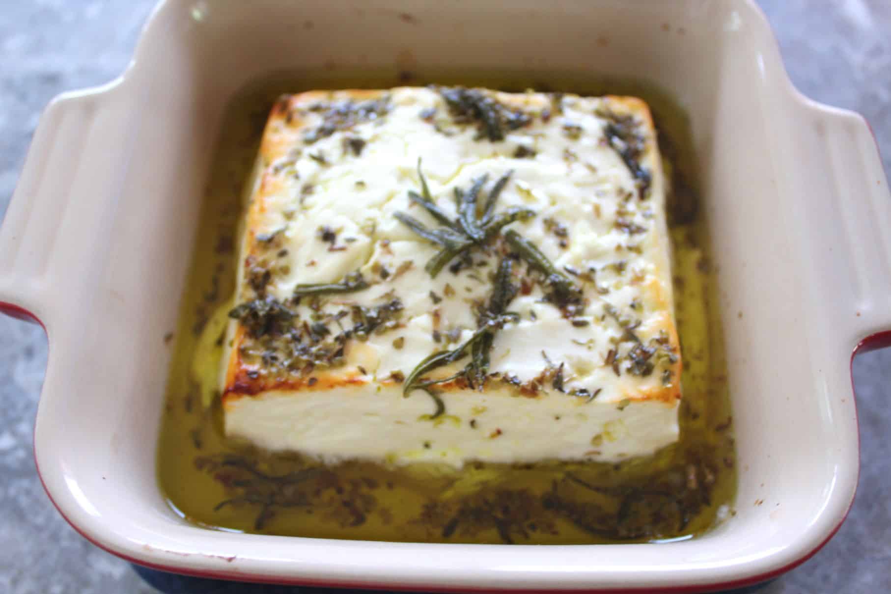 A baking dish showing a freshly baked block of feta cheese in olive oil and covered in herbs.