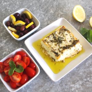 Baked feta cheese in olive oil and herbs shown next to two other sides, olives with lemon and grape tomatoes.