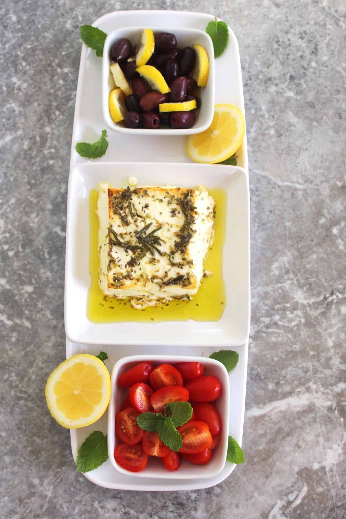 Appetizer tray with 3 separate side dishes: from top to bottom, olives with lemon, baked feta cheese, herbed in olive oil and lastly grape tomatoes with mint.