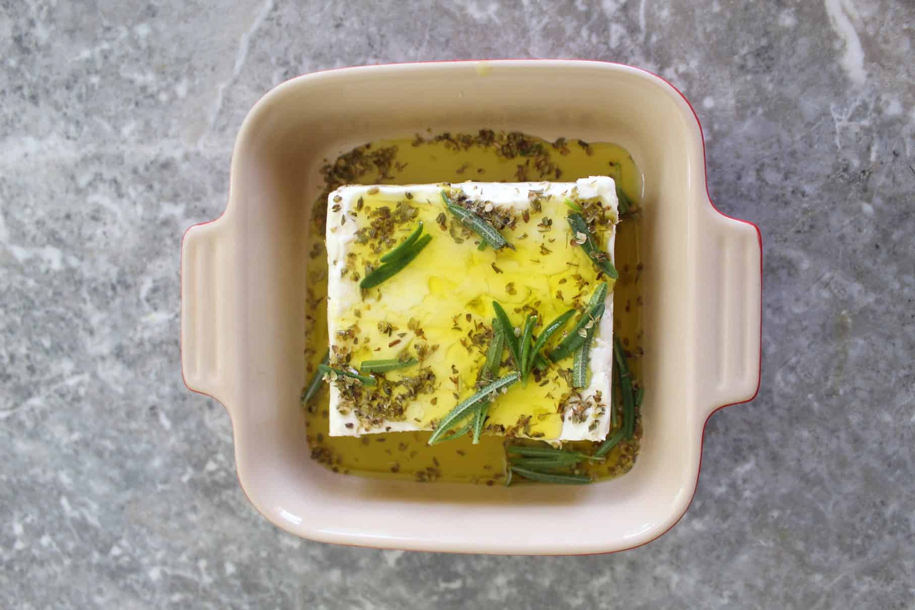 A square baking dish with a block of feta cheese, covered in olive oil and herbs. 