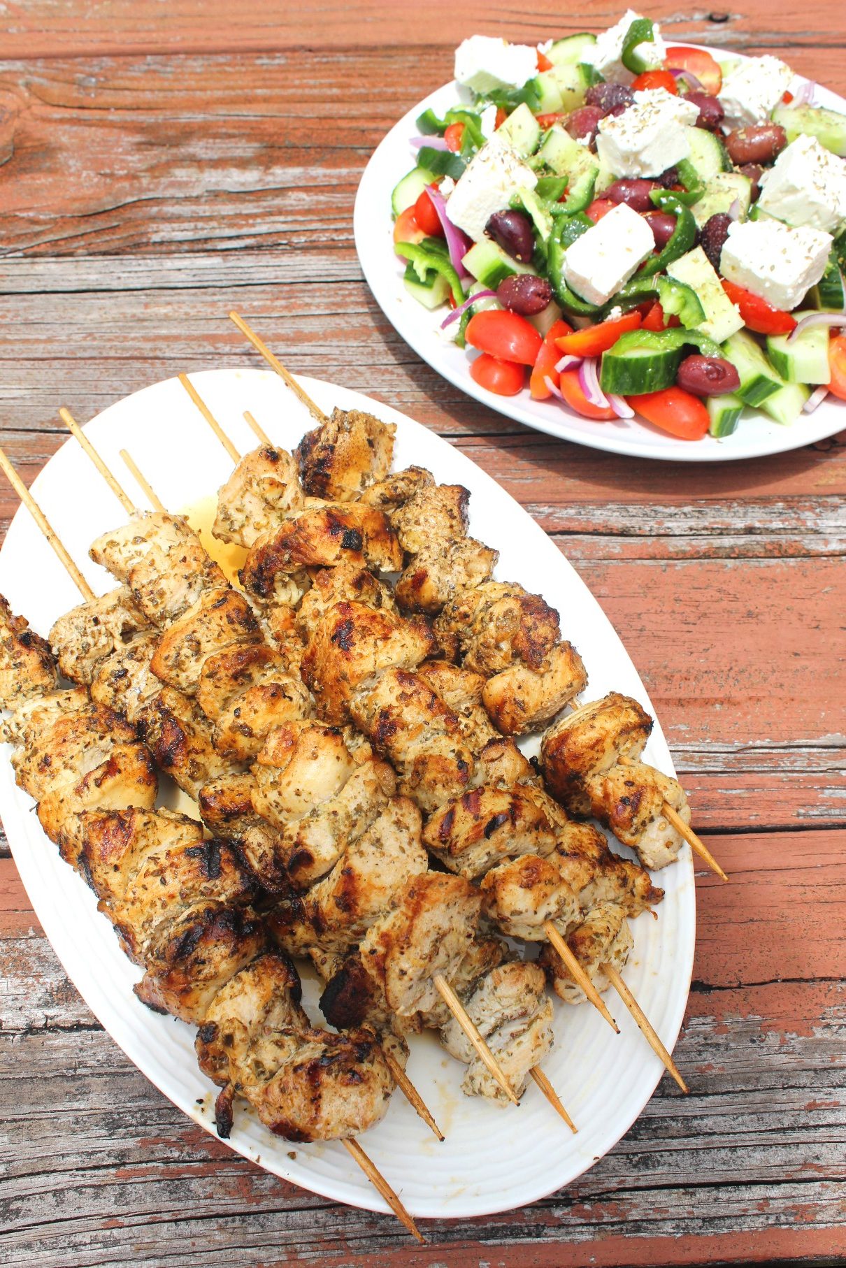 An oval platter of chicken kabobs still in skewers next to a plate of greek salad. Both plates are on a rustic looking wood surface.