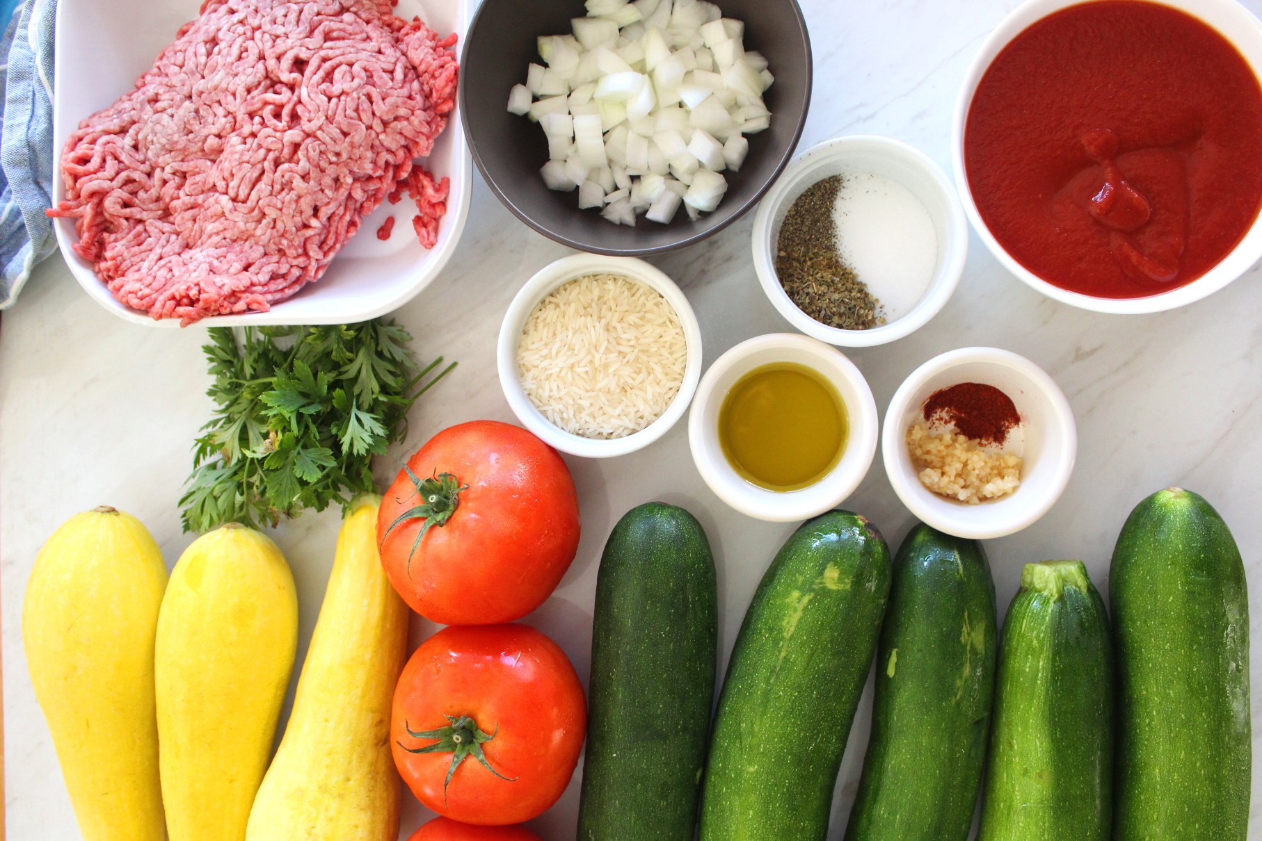Picture shows most of the ingredients used for the Mediterranean baked stuffed zucchini casserole. You can see zuchinis, squash, tomatoes, tomato sauce, onions, spices, rice, oil, and ground beef. 