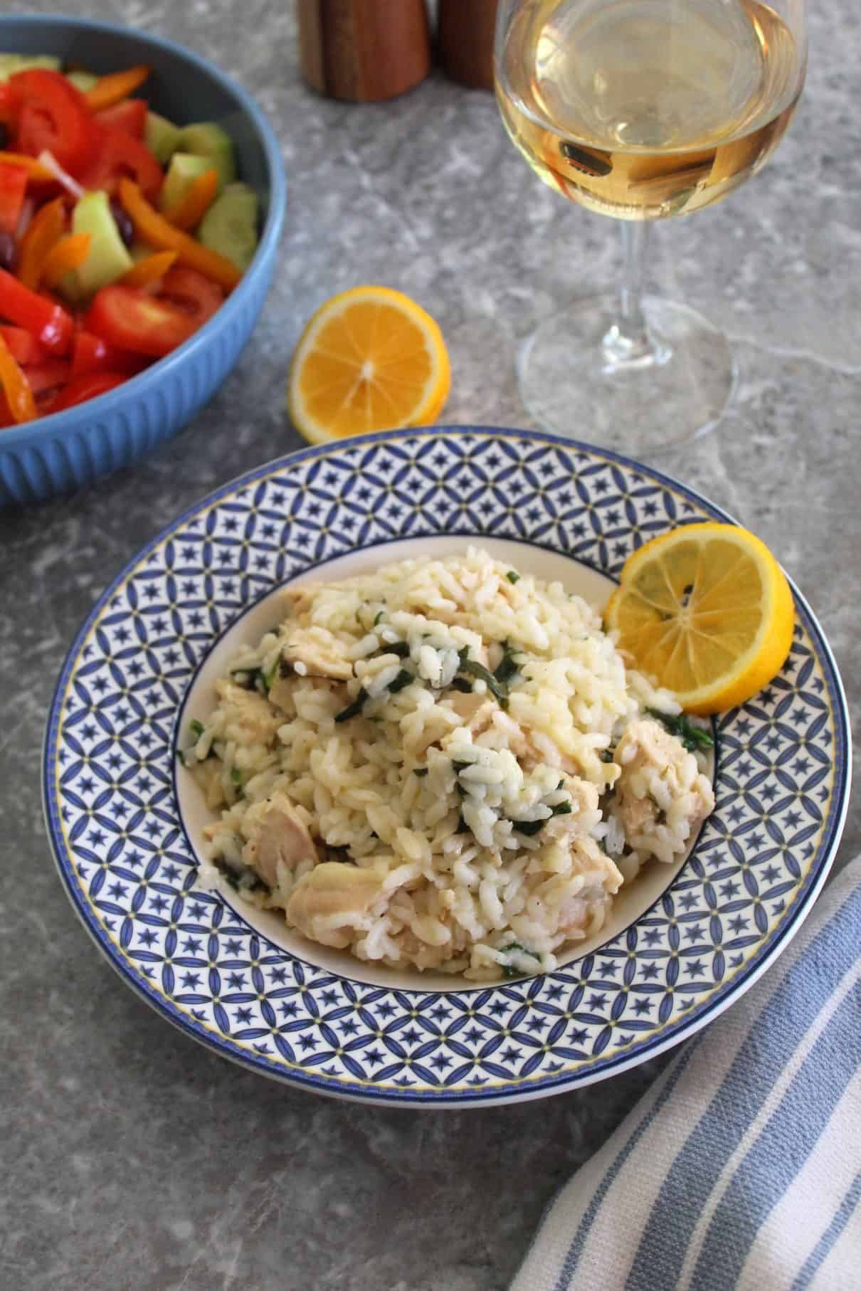 A plate of risotto garnished with lemon. Risotto seems to have chicken and spinach in it. Out of focus you see a salad bowl, a half lemon and a glass of rose wine.