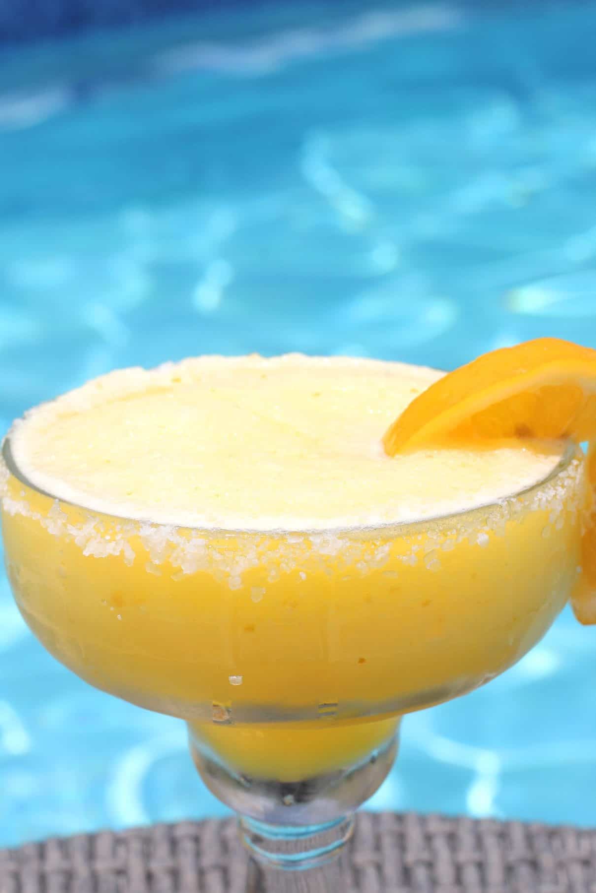 A frozen margarita next to the pool. Cup has a salty rim and a slice of Meyer Lemon on the side. Cup also looks like it has a condensed outer part. 