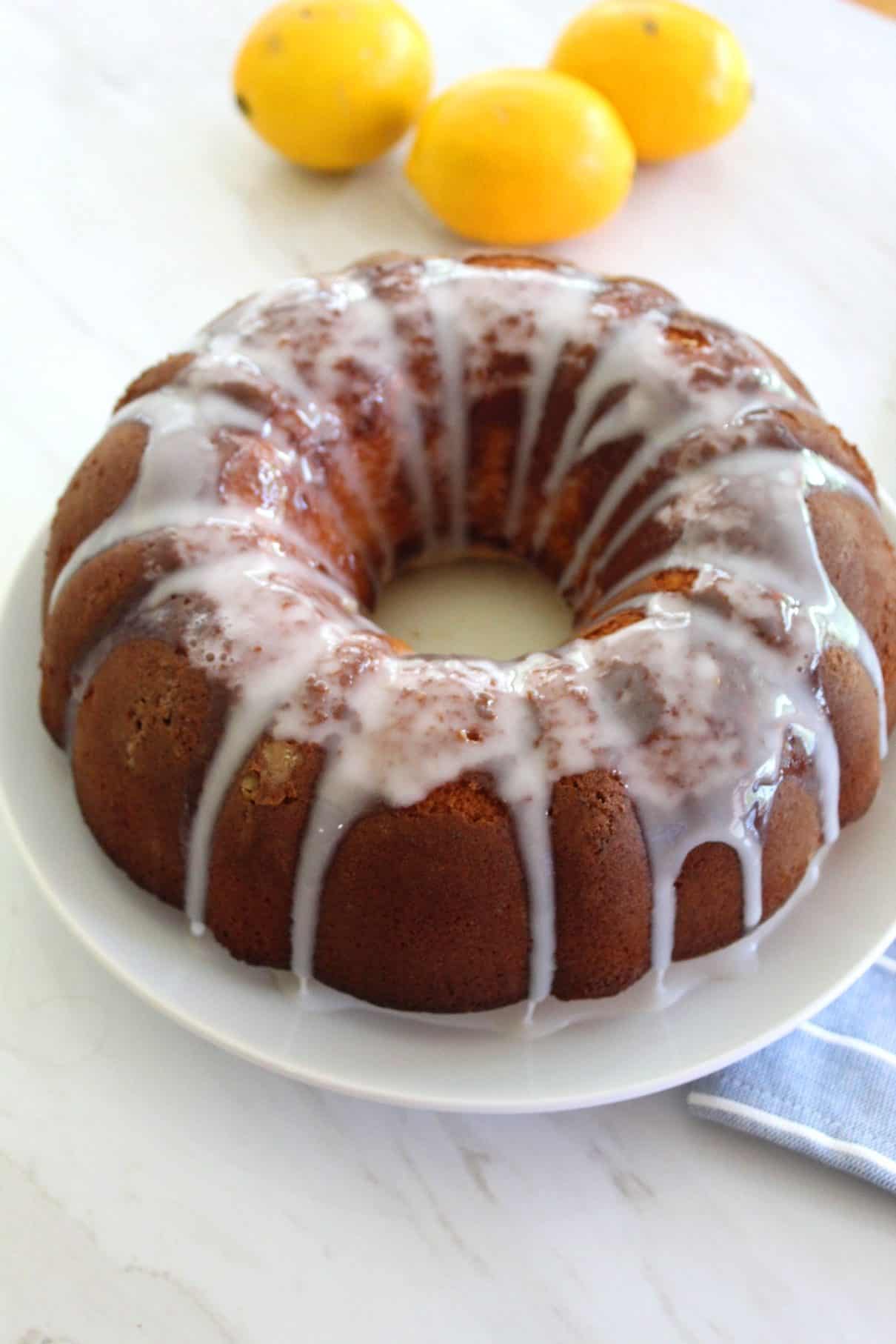A round platter showing a Meyer Lemon bundt  cake with fresh icing dripping down on the sides. Fresh lemons are shown in the background.