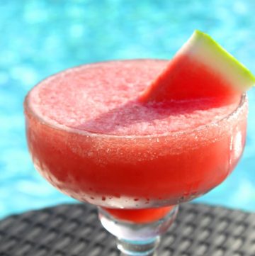 A frozen red drink in a margarita cup, garnished with a watermelon slice. Cup is placed in a table by the pool.