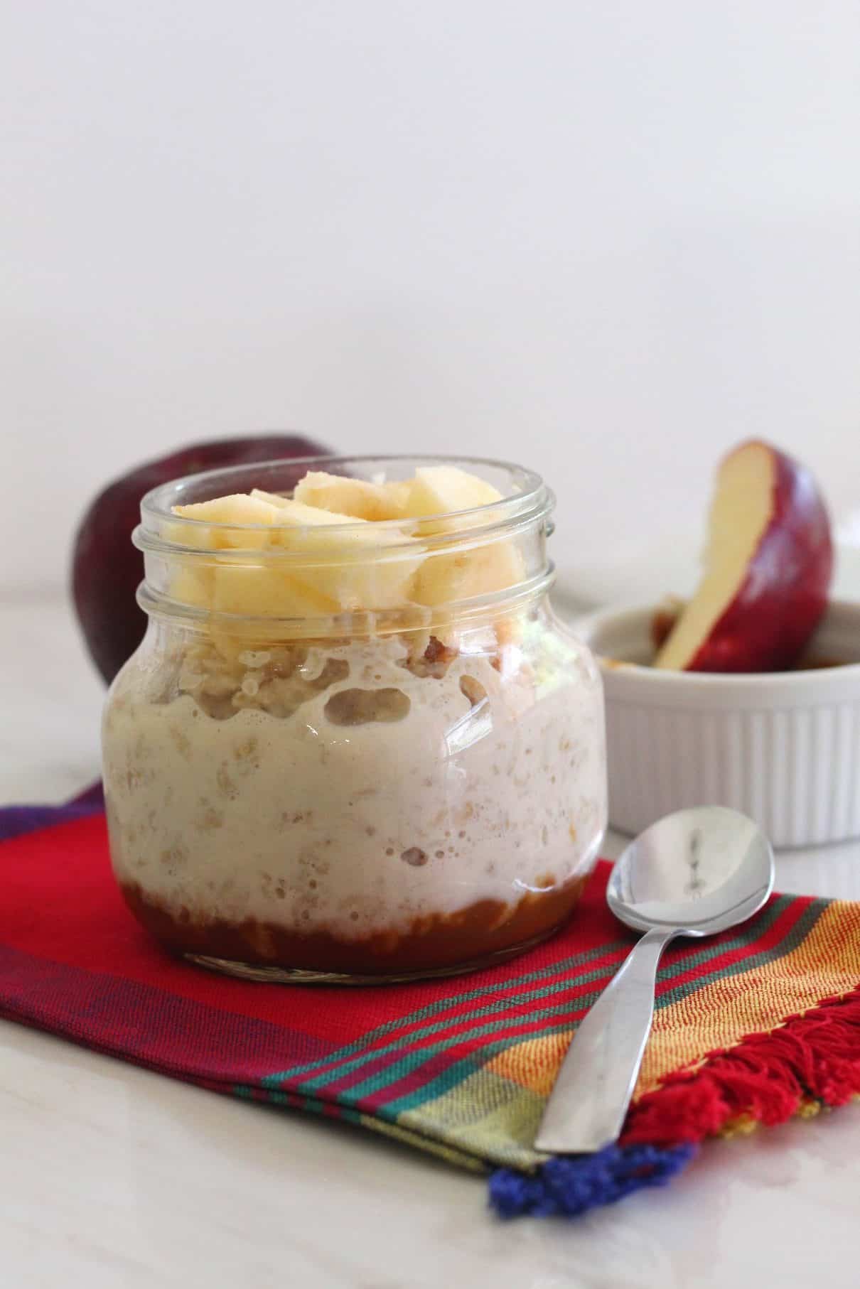 An oats jar topped with apples, dulce de leche is seen at the bottom of the jar. There's also an apple in the background and a slice of apple dipped in dulce de leche on the other side. 