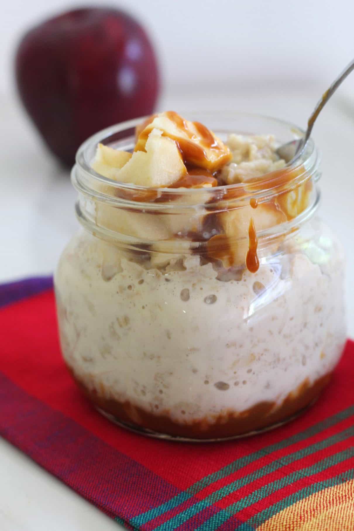 Creamy oatmeal topped with crisp apples and dulce de leche, ready to be dug in. Breakfast jar is placed over a red napkin and spoon is already in the jar. There's a little drizzle of dulce de leche dripping on the outside of the jar. 