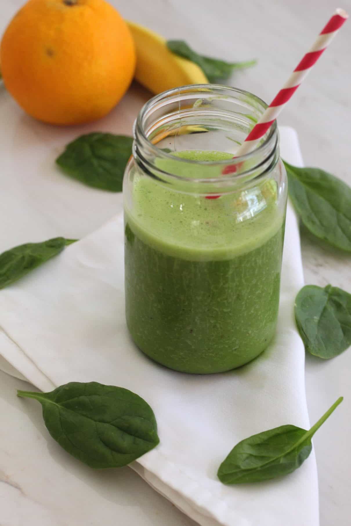 A smoothie jar with a paper straw. Smoothie is green. You can see an orange and a banana in the background as well as scattered spinach leaves.
