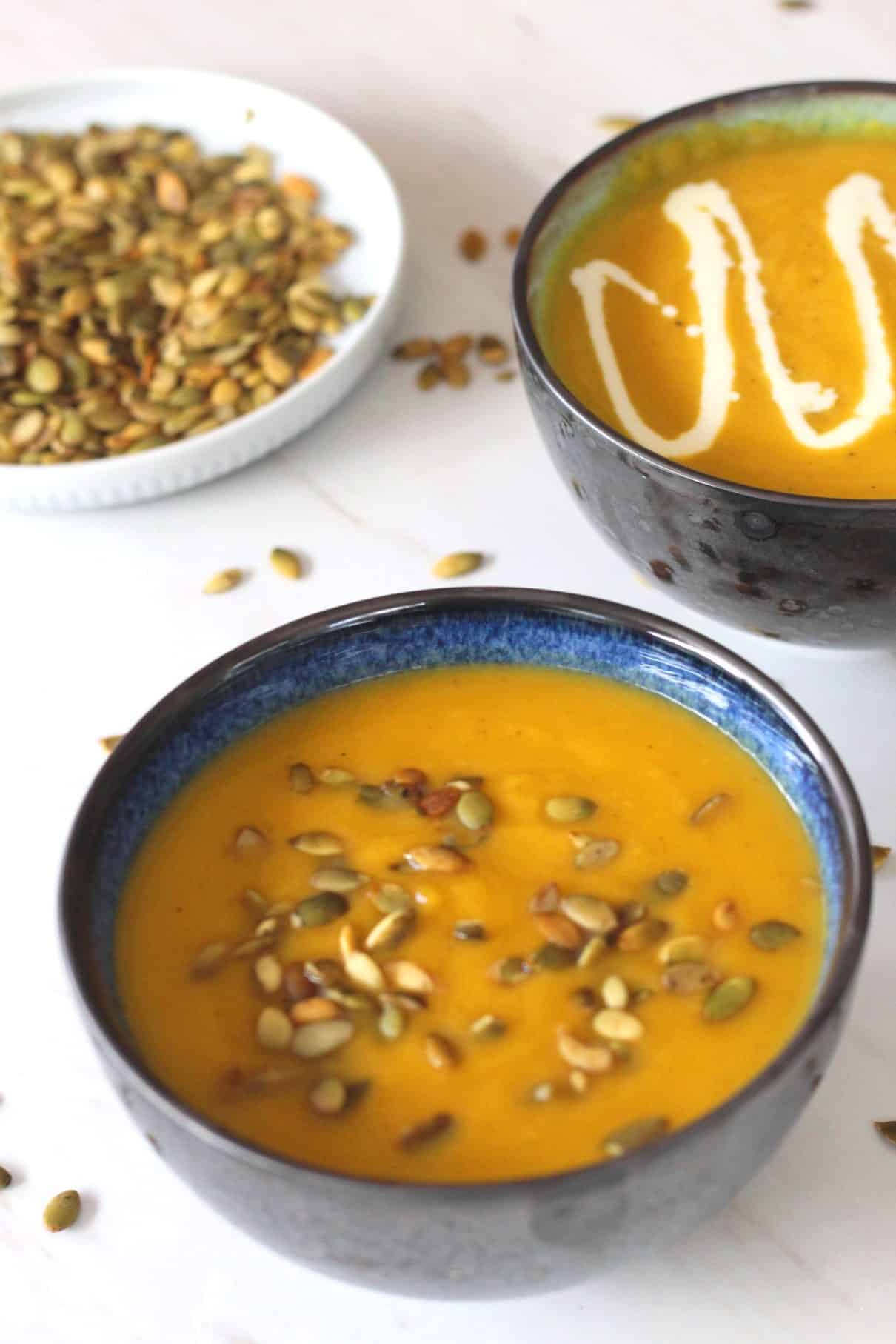 A bowl of soup forefront shown with a creamy Fall soup. Soup is topped with pumpkin seeds. There's another bowl of soup in the background that is topped with cream. 