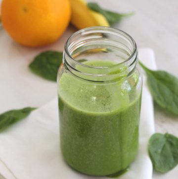 A jar with green smoothie next to spinach leaves, an orange and a banana