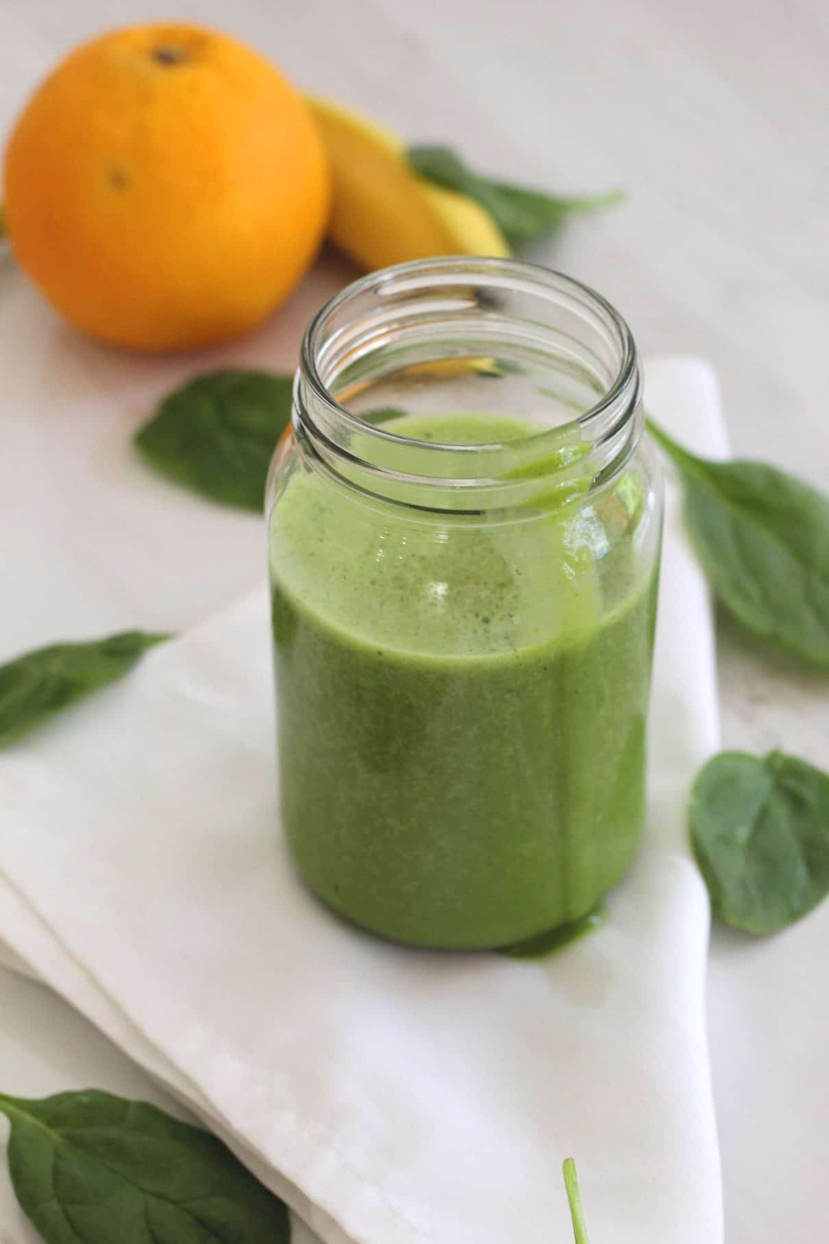 Green smoothie with spinach, orange, banana and oat milk. Jar with smoothie is shown over a white napkin with a trail of smoothie spilling over the side of the jar.