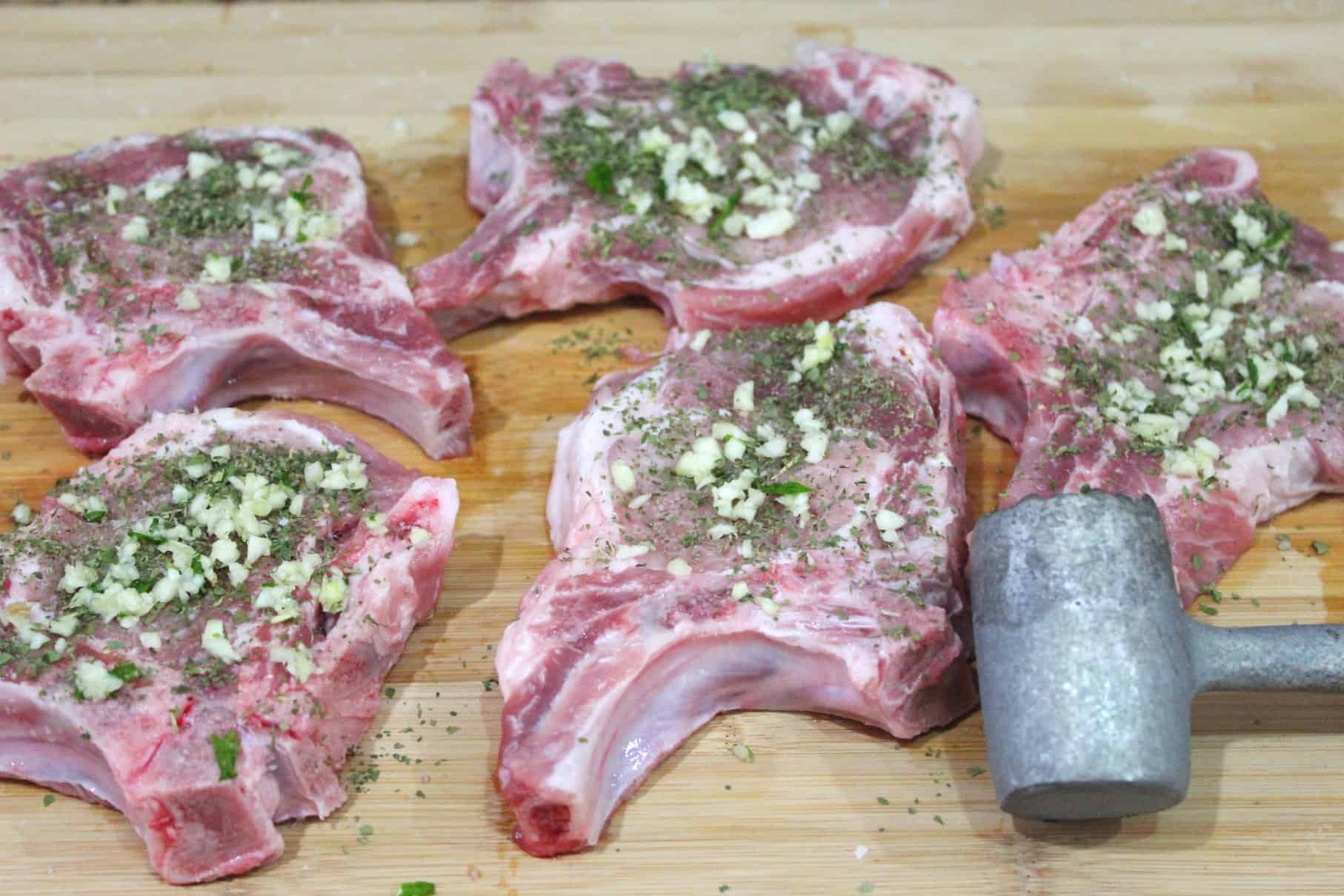 A wooden block with pork chops seasoned with herbs and garlic. Chops are shown next to a meat tenderizer hammer. 