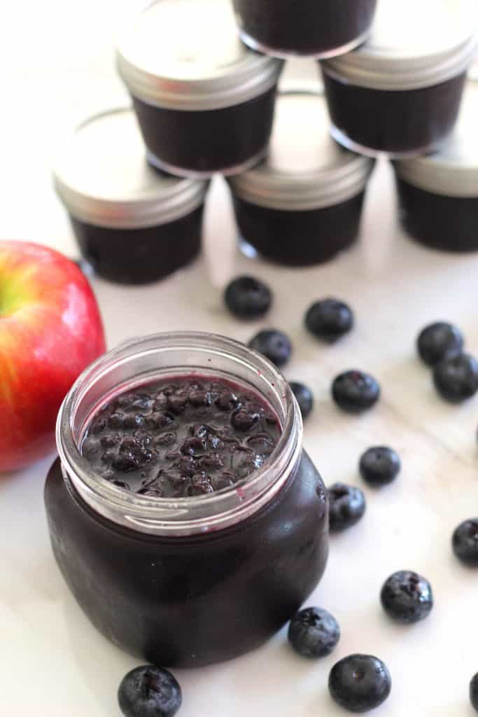 Jar of blueberry jam at forefront surrounded by fresh blueberries. In the background you see an apple and several jars of jam.