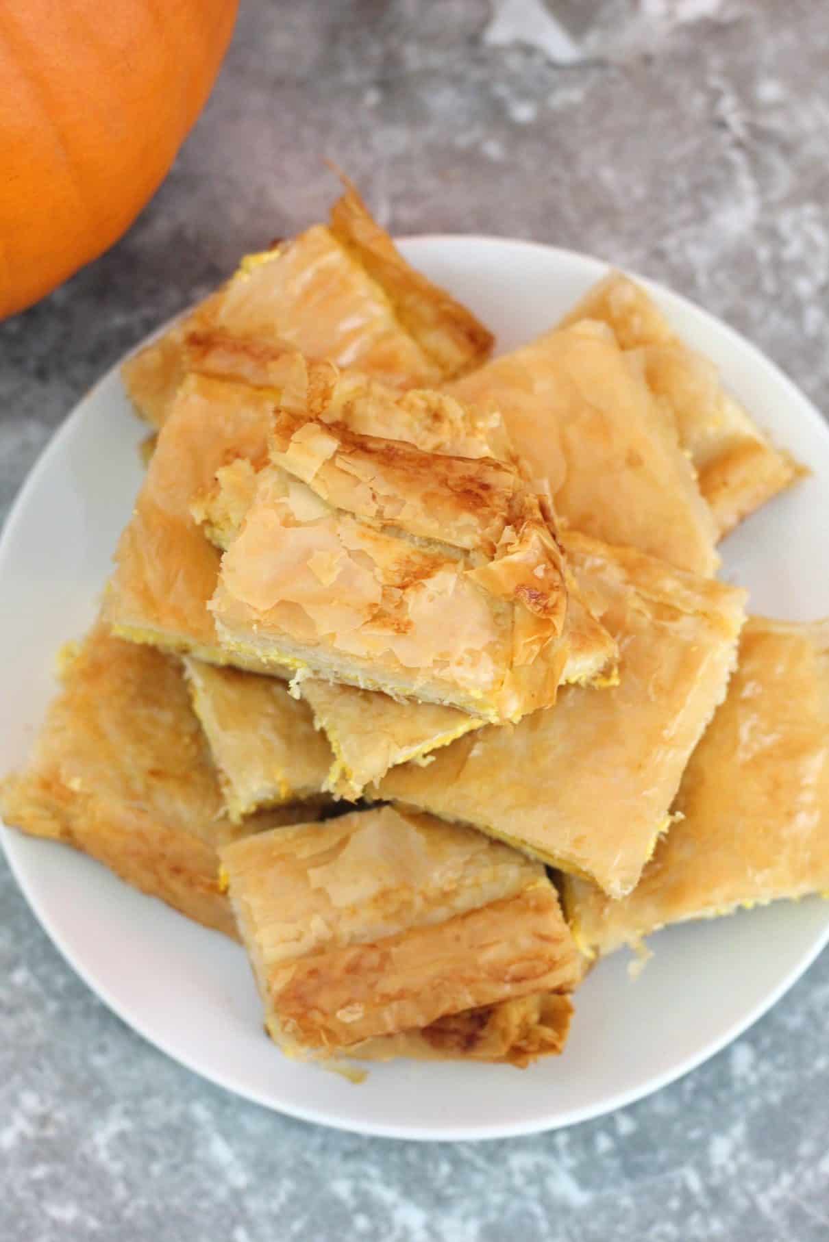 A plate full of slices of byrek, a phyllo pumpkin pie. In the background you can see a little bit of a pumpkin.