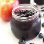 A jar of Blueberry Apple Jam, shown next to fresh blueberries and an apple.